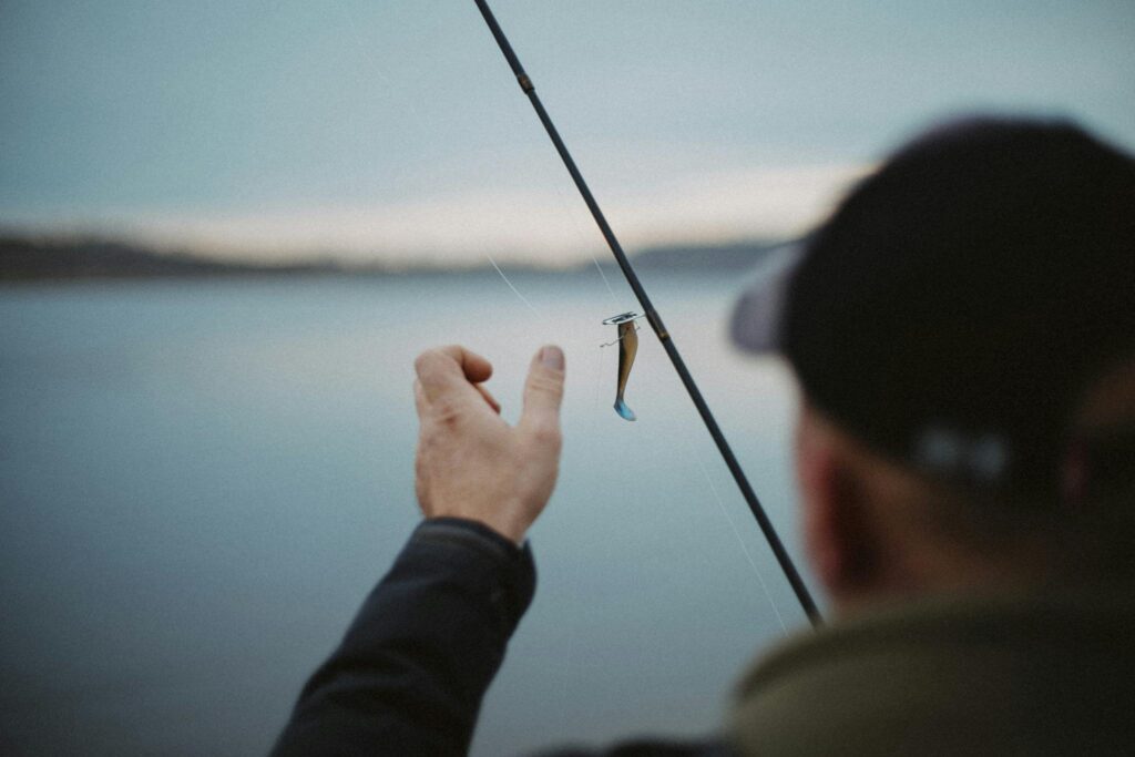 A fisherman adjusts his lure on a fishing rod by a calm lake, creating a peaceful outdoor scene.