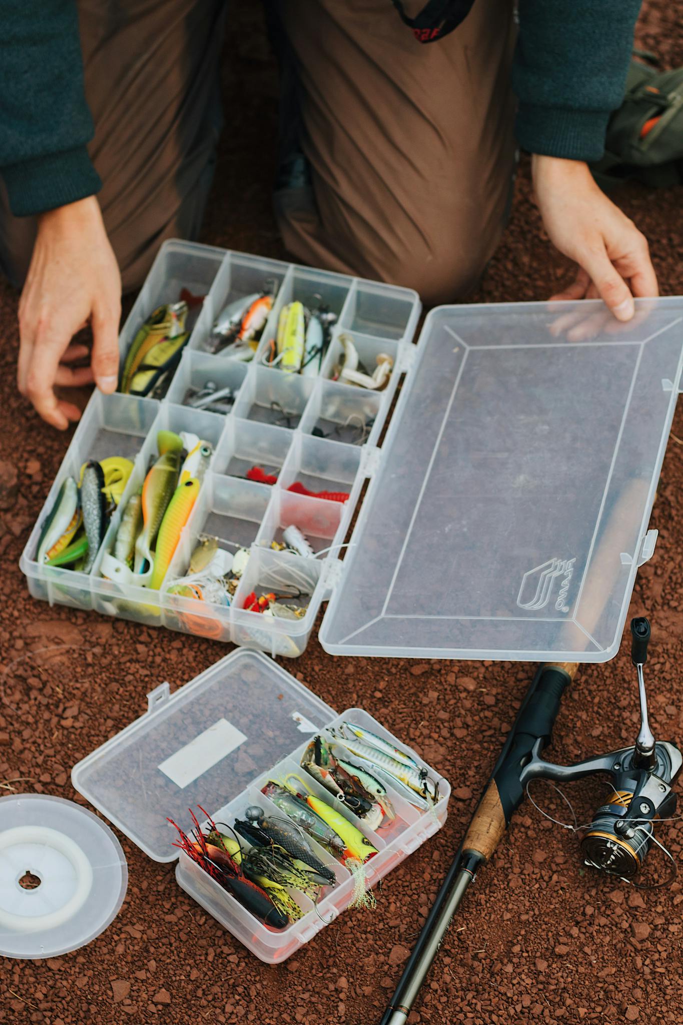 Fisherman arranging fishing lures in a tackle box beside a fishing rod.