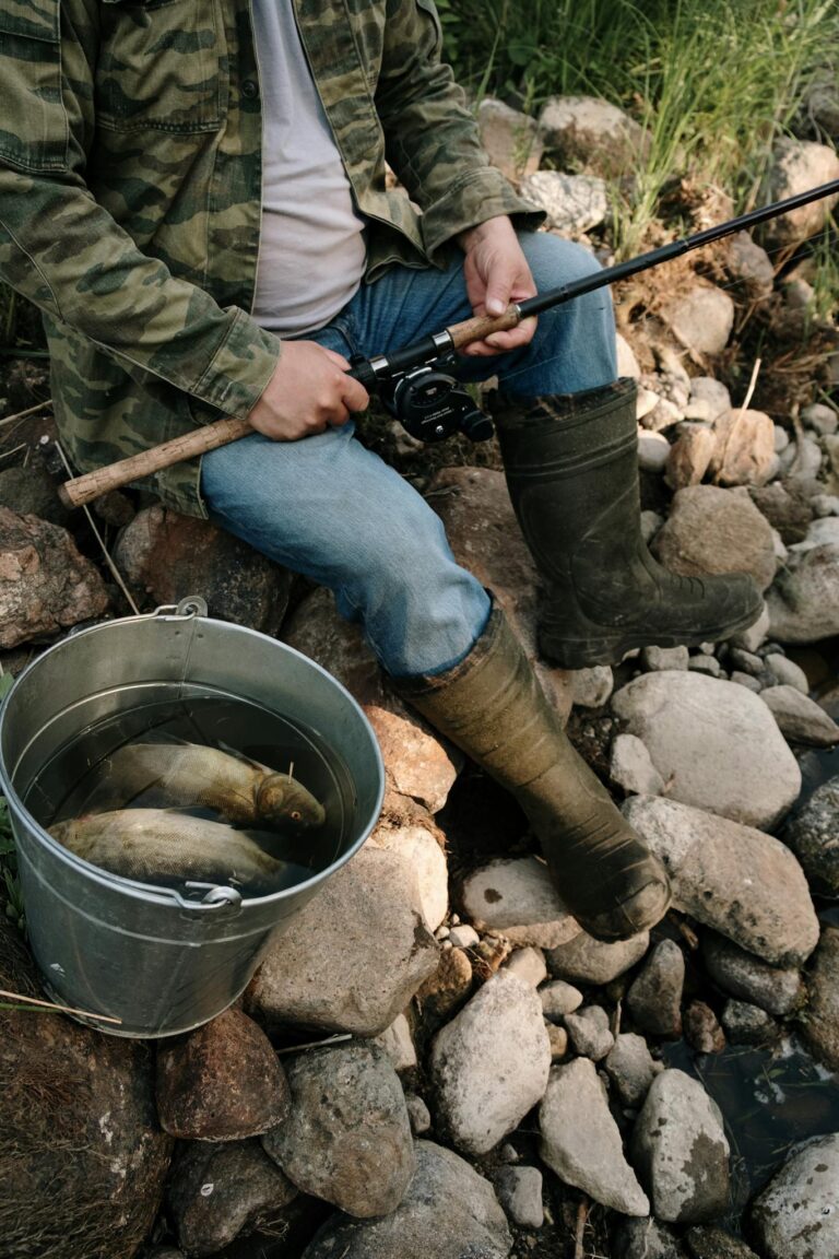 Fisherman sitting on rocks holding a rod, a bucket with fish beside him, wearing camouflage and boots.